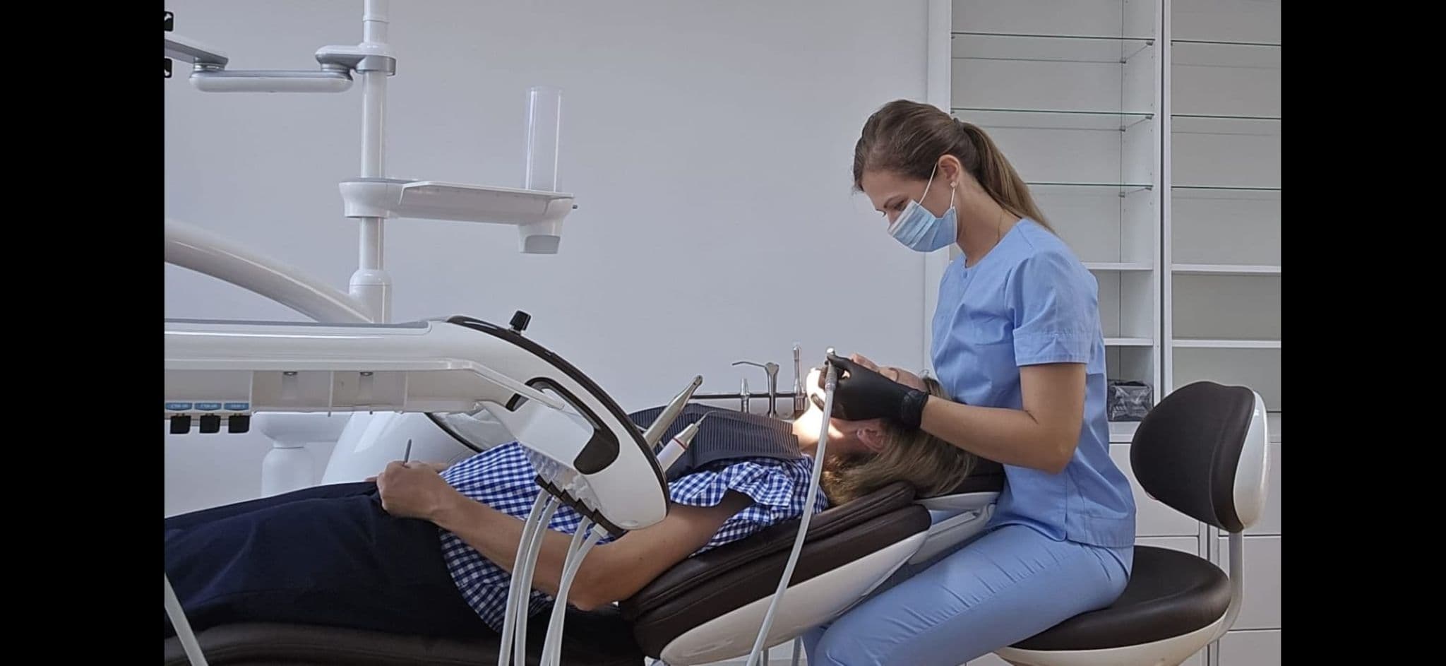 Dentist in blue scrubs and mask treats a patient lying in a modern dental chair.