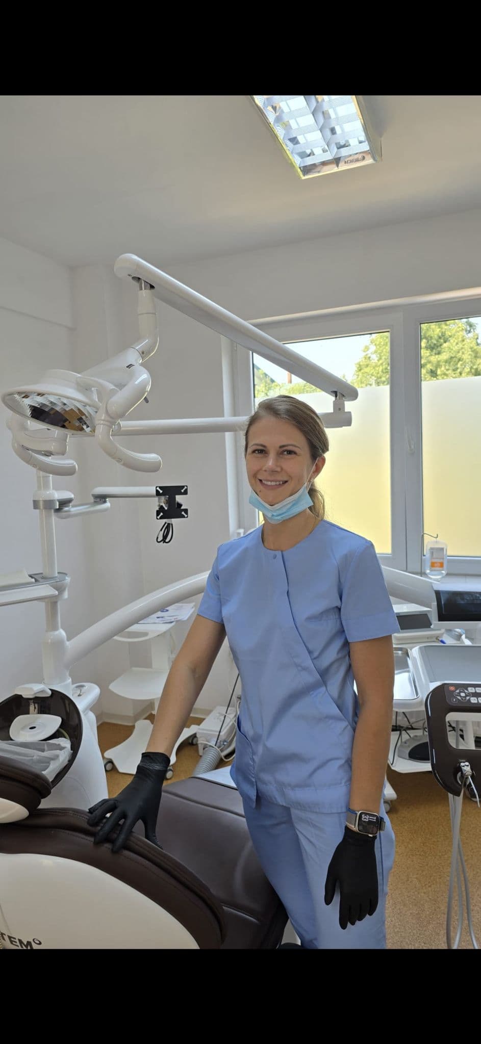 Smiling female dentist in light blue scrubs and black gloves standing in a dental office.
