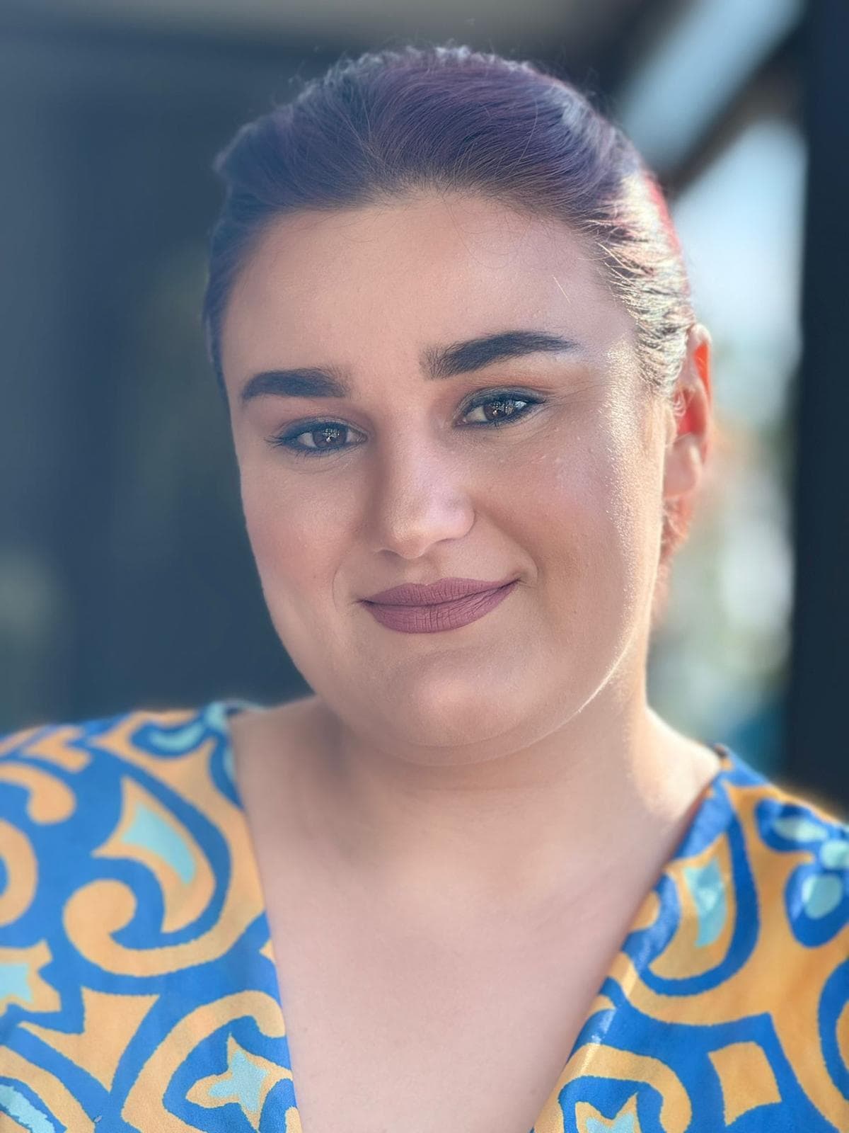 Smiling woman with purple hair and dark eyebrows wearing a blue and yellow patterned top.